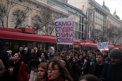 Πλήθος ανθρώπων και διαδηλωτές σε δρόμο με κόκκινα οχήματα και πλακάτ.