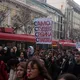 Πλήθος ανθρώπων και διαδηλωτές σε δρόμο με κόκκινα οχήματα και πλακάτ.