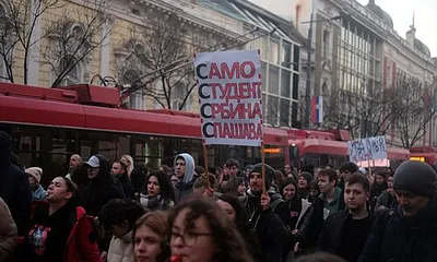 Πλήθος ανθρώπων και διαδηλωτές σε δρόμο με κόκκινα οχήματα και πλακάτ.