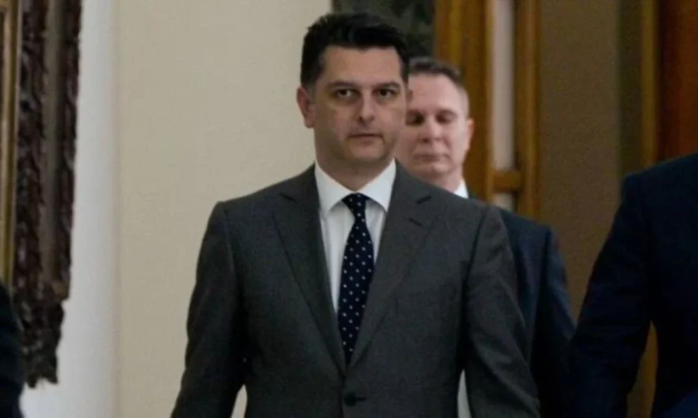 Άντρας με κοστούμι και γραβάτα περπατά σε εσωτερικό χώρο με άλλα πρόσωπα στο βάθος.