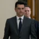 Άντρας με κοστούμι και γραβάτα περπατά σε εσωτερικό χώρο με άλλα πρόσωπα στο βάθος.