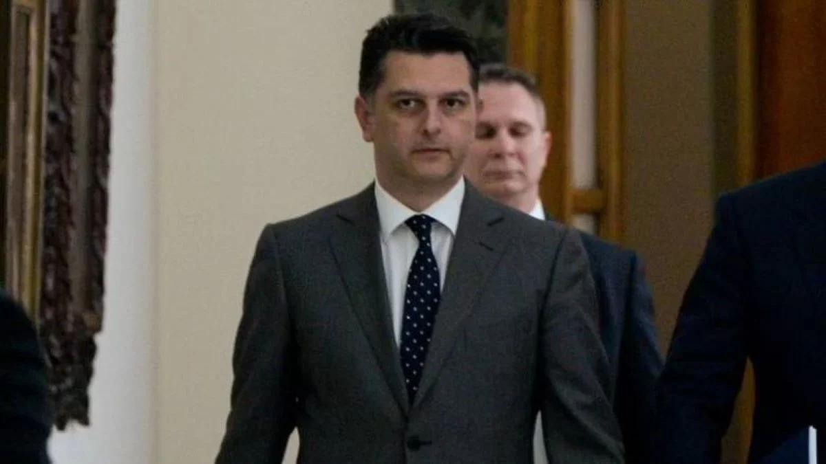 Άντρας με κοστούμι και γραβάτα περπατά σε εσωτερικό χώρο με άλλα πρόσωπα στο βάθος.