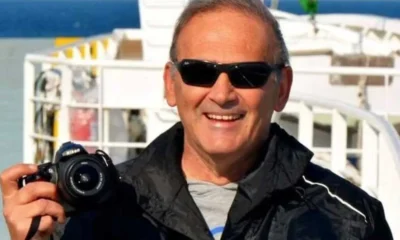 Άντρας φοράει μαύρα γυαλιά ηλίου και κρατά φωτογραφική μηχανή σε εξωτερικό χώρο.