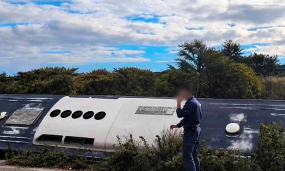 Όρθιος άνδρας με σκούρα στολή δίπλα σε ανατροπή οχήματος σε εξωτερικό χώρο με θάμνους και συννεφιασμένο ουρανό.