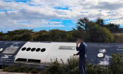 Όρθιος άνδρας με σκούρα στολή δίπλα σε ανατροπή οχήματος σε εξωτερικό χώρο με θάμνους και συννεφιασμένο ουρανό.