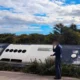 Όρθιος άνδρας με σκούρα στολή δίπλα σε ανατροπή οχήματος σε εξωτερικό χώρο με θάμνους και συννεφιασμένο ουρανό.