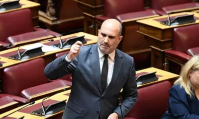 Άντρας με κοστούμι μιλάει σε αίθουσα με κόκκινες πολυθρόνες και έδρανα