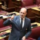 Άντρας με κοστούμι μιλάει σε αίθουσα με κόκκινες πολυθρόνες και έδρανα