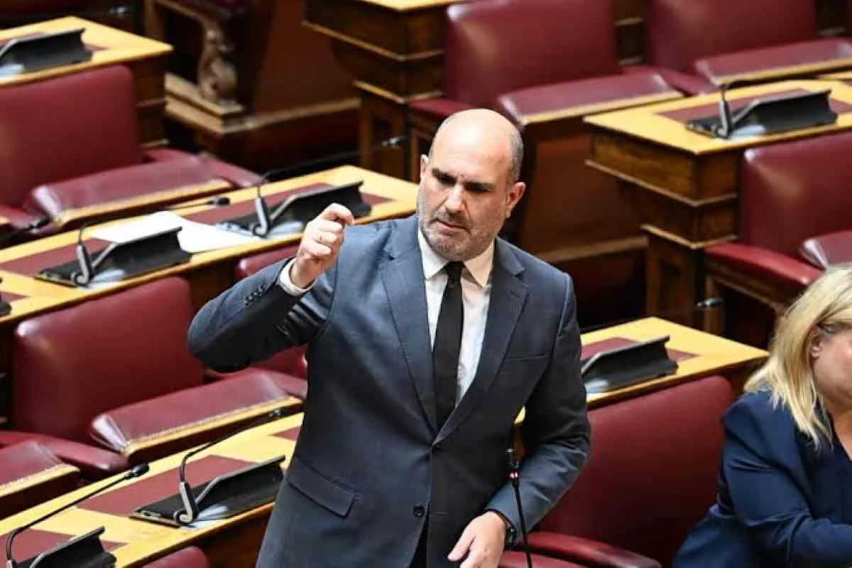 Άντρας με κοστούμι μιλάει σε αίθουσα με κόκκινες πολυθρόνες και έδρανα