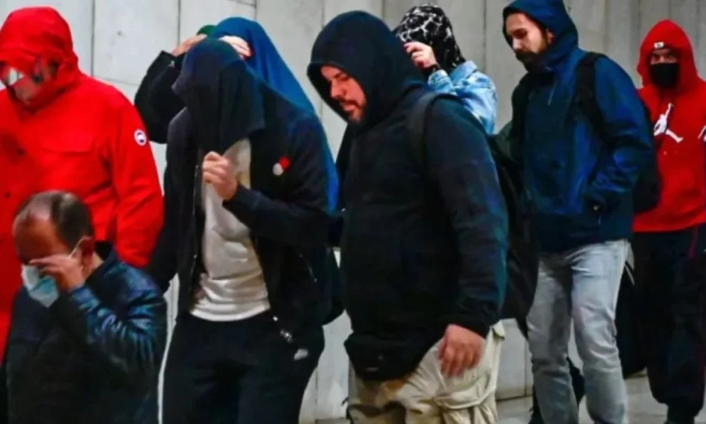 Ομάδα ατόμων με κουκούλες και μπουφάν βαδίζει σε εσωτερικό χώρο με ανοιχτόχρωμο τοίχο.