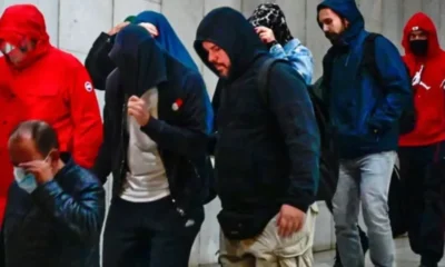 Ομάδα ατόμων με κουκούλες και μπουφάν βαδίζει σε εσωτερικό χώρο με ανοιχτόχρωμο τοίχο.