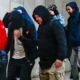 Ομάδα ατόμων με κουκούλες και μπουφάν βαδίζει σε εσωτερικό χώρο με ανοιχτόχρωμο τοίχο.