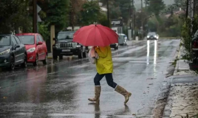 Άτομο με κόκκινη ομπρέλα, κίτρινο αδιάβροχο και μπότες διασχίζει βρεγμένο δρόμο σε περιοχή με παρκαρισμένα αυτοκίνητα