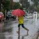 Άτομο με κόκκινη ομπρέλα, κίτρινο αδιάβροχο και μπότες διασχίζει βρεγμένο δρόμο σε περιοχή με παρκαρισμένα αυτοκίνητα