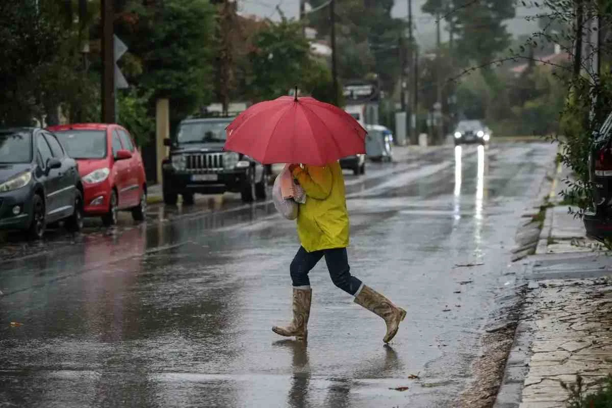 Άτομο με κόκκινη ομπρέλα, κίτρινο αδιάβροχο και μπότες διασχίζει βρεγμένο δρόμο σε περιοχή με παρκαρισμένα αυτοκίνητα