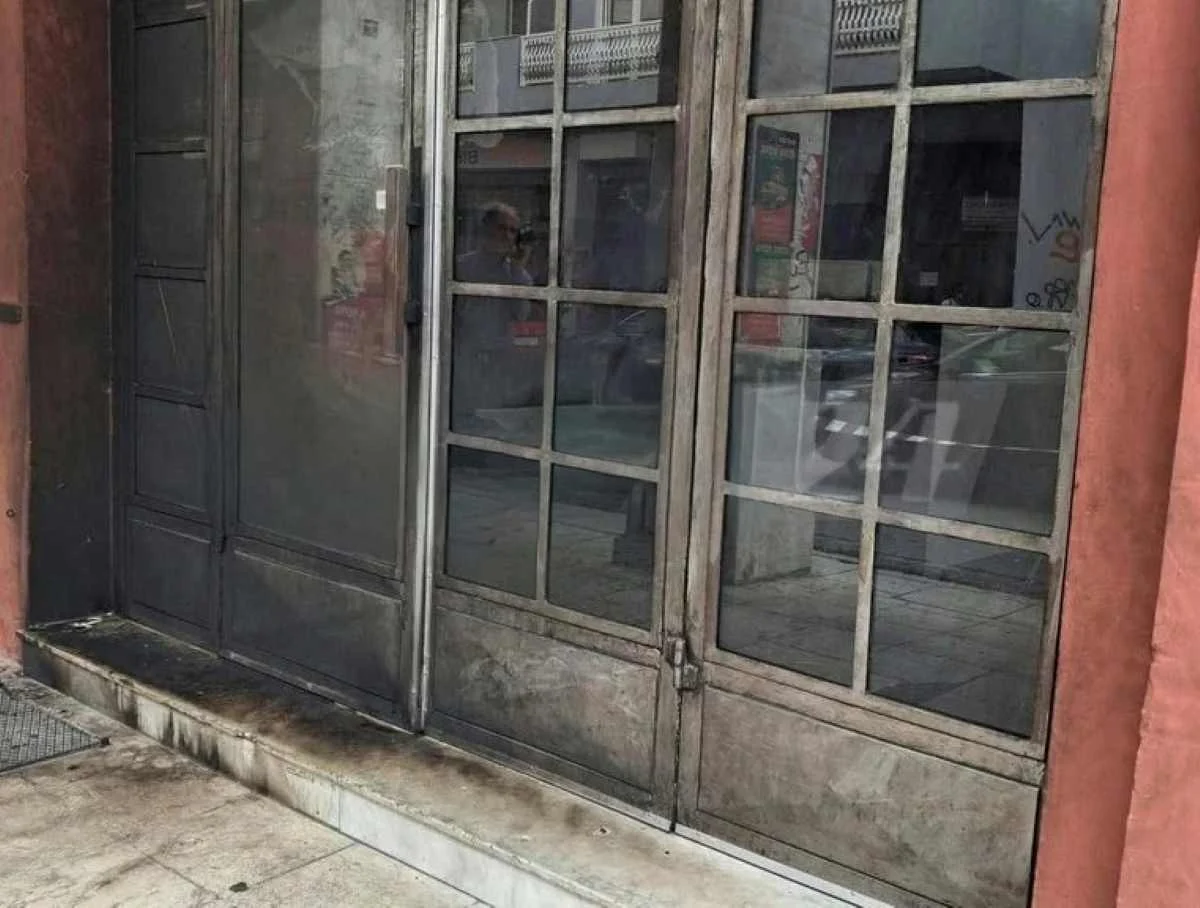 Η είσοδος καταστήματος με μεταλλική πόρτα και γυάλινα πλαίσια, με μαύρα σηγματα στην επιφάνεια του δαπέδου.