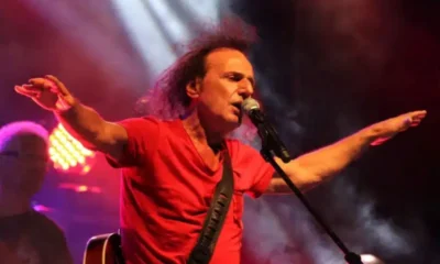 Άντρας με κόκκινη μπλούζα τραγουδάει σε μικρόφωνο πάνω σε σκηνή με φωτισμό και καπνό.