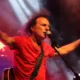 Άντρας με κόκκινη μπλούζα τραγουδάει σε μικρόφωνο πάνω σε σκηνή με φωτισμό και καπνό.