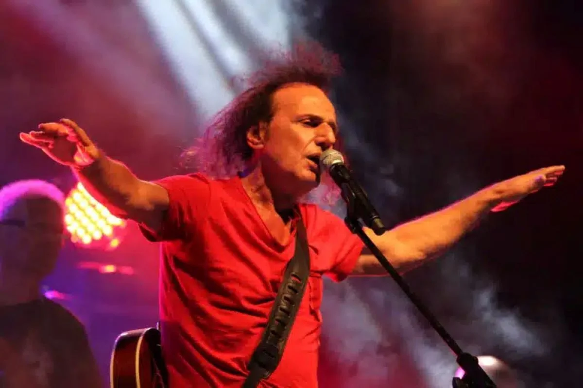 Άντρας με κόκκινη μπλούζα τραγουδάει σε μικρόφωνο πάνω σε σκηνή με φωτισμό και καπνό.