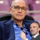 Δύο άνδρες ποζάρουν σε φωτογραφία, ο ένας φορά γυαλιά και μπλε σακάκι με πουκάμισο και ο άλλος κοστούμι με γραβάτα.