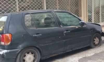 Ένα παλιό σκούρο αυτοκίνητο είναι σταθμευμένο στο δρόμο μπροστά από κτίριο με κλειστά ρολά, έχοντας σκονισμένη όψη.