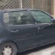 Ένα παλιό σκούρο αυτοκίνητο είναι σταθμευμένο στο δρόμο μπροστά από κτίριο με κλειστά ρολά, έχοντας σκονισμένη όψη.