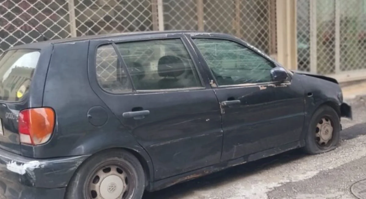 Ένα παλιό σκούρο αυτοκίνητο είναι σταθμευμένο στο δρόμο μπροστά από κτίριο με κλειστά ρολά, έχοντας σκονισμένη όψη.