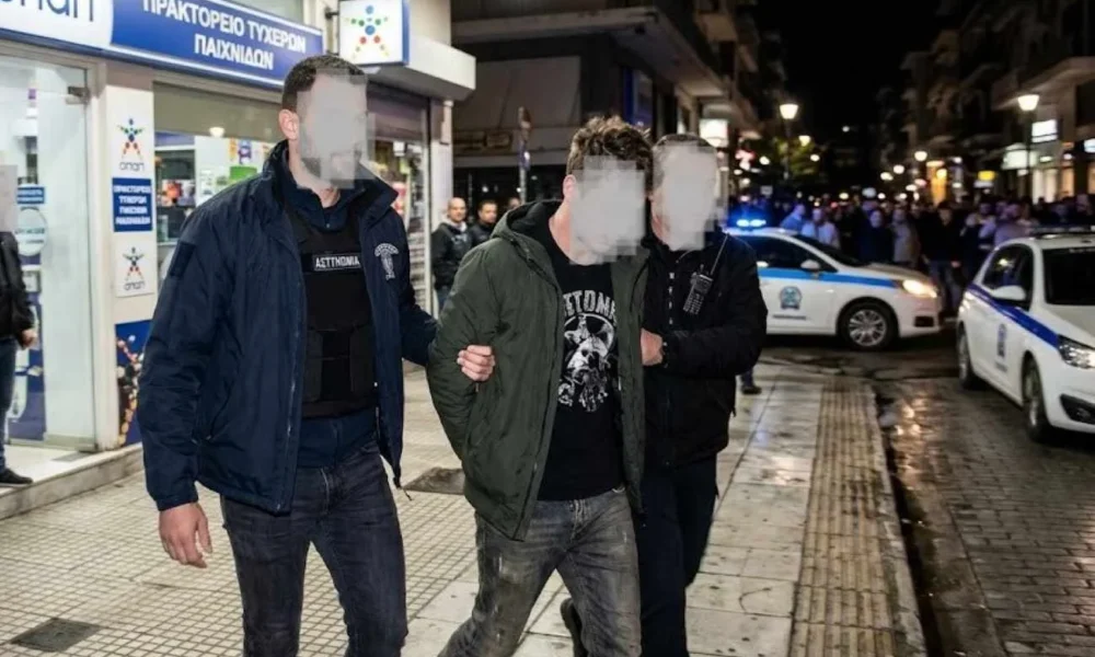 Άνδρας με χειροπέδες οδηγείται από αστυνομικούς σε δρόμο μπροστά από κατάστημα και περιπολικό