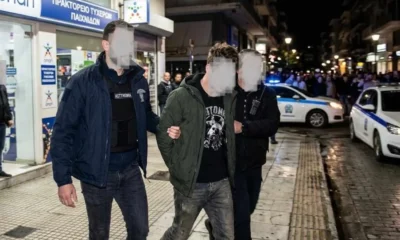 Άνδρας με χειροπέδες οδηγείται από αστυνομικούς σε δρόμο μπροστά από κατάστημα και περιπολικό