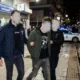 Άνδρας με χειροπέδες οδηγείται από αστυνομικούς σε δρόμο μπροστά από κατάστημα και περιπολικό