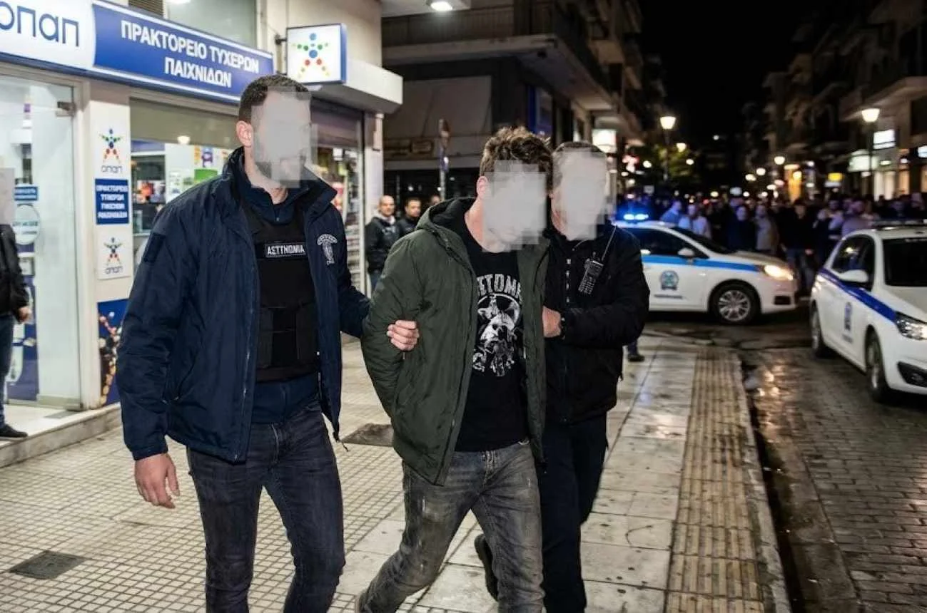 Άνδρας με χειροπέδες οδηγείται από αστυνομικούς σε δρόμο μπροστά από κατάστημα και περιπολικό