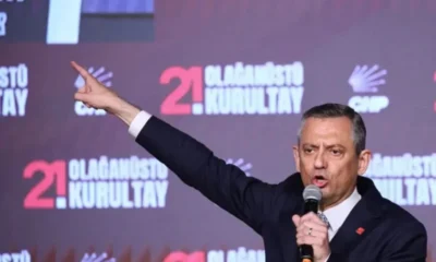 Άνδρας με κοστούμι κρατάει μικρόφωνο και δείχνει με το χέρι του μπροστά σε τοίχο με επιγραφές.
