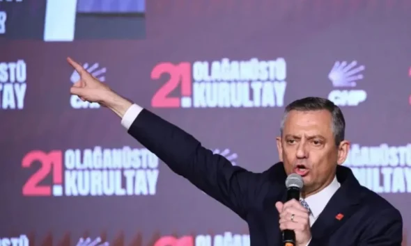 Άνδρας με κοστούμι κρατάει μικρόφωνο και δείχνει με το χέρι του μπροστά σε τοίχο με επιγραφές.
