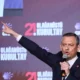 Άνδρας με κοστούμι κρατάει μικρόφωνο και δείχνει με το χέρι του μπροστά σε τοίχο με επιγραφές.