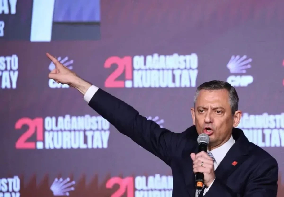 Άνδρας με κοστούμι κρατάει μικρόφωνο και δείχνει με το χέρι του μπροστά σε τοίχο με επιγραφές.