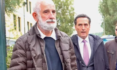 Δύο άνδρες περπατούν σε εξωτερικό χώρο, ο ένας με λευκή γενειάδα και μπουφάν, και ο άλλος με κοστούμι και γραβάτα.