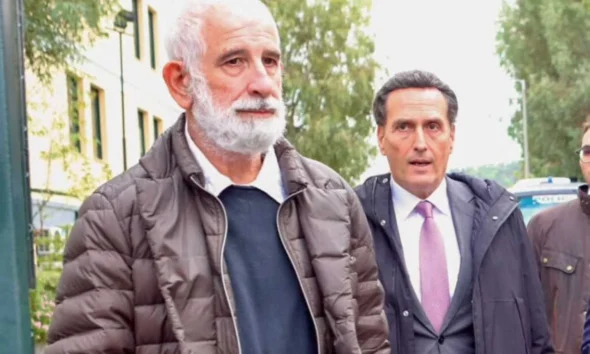 Δύο άνδρες περπατούν σε εξωτερικό χώρο, ο ένας με λευκή γενειάδα και μπουφάν, και ο άλλος με κοστούμι και γραβάτα.