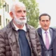 Δύο άνδρες περπατούν σε εξωτερικό χώρο, ο ένας με λευκή γενειάδα και μπουφάν, και ο άλλος με κοστούμι και γραβάτα.
