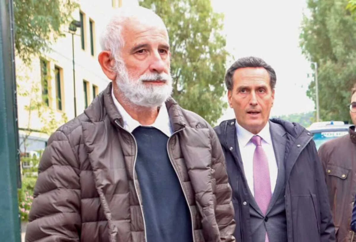 Δύο άνδρες περπατούν σε εξωτερικό χώρο, ο ένας με λευκή γενειάδα και μπουφάν, και ο άλλος με κοστούμι και γραβάτα.