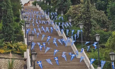 Υπαίθρια σκάλα στολισμένη με μικρές ελληνικές σημαίες κατά μήκος της, ανάμεσα σε πράσινα δέντρα και κολώνες φωτισμού.