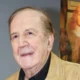Πορτραίτο ηλικιωμένου άνδρα με ανοιχτόχρωμο σακάκι και σκούρο πουκάμισο, με πίνακα ζωγραφικής στο βάθος