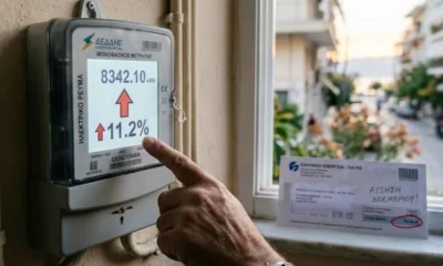 Χέρι αγγίζει ψηφιακό μετρητή ρεύματος που δείχνει κόκκινο βέλος και ποσοστό ανόδου, δίπλα σε λογαριασμό.