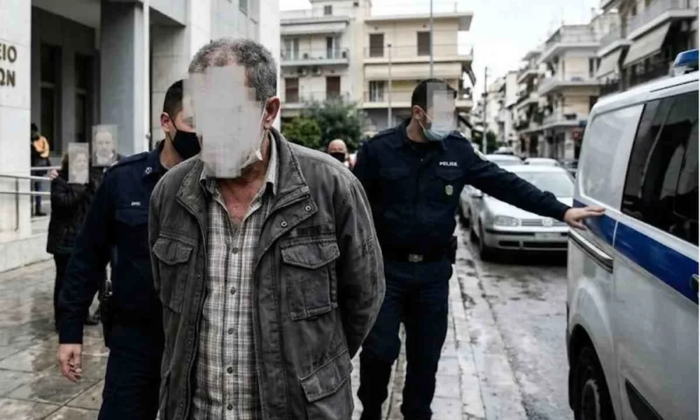 Άνδρας με καλυμμένα χαρακτηριστικά συνοδεύεται από αστυνομικούς κοντά σε όχημα.