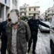 Άνδρας με καλυμμένα χαρακτηριστικά συνοδεύεται από αστυνομικούς κοντά σε όχημα.