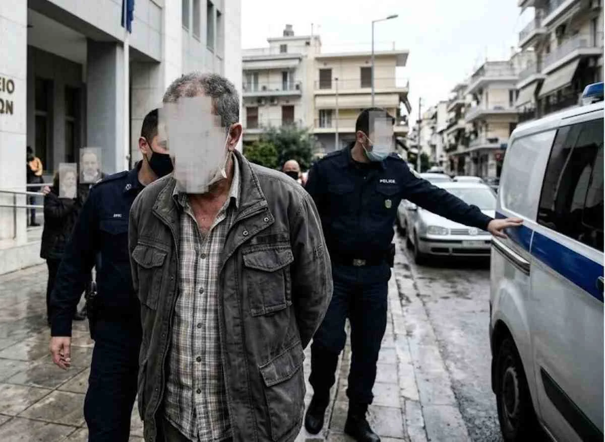 Άνδρας με καλυμμένα χαρακτηριστικά συνοδεύεται από αστυνομικούς κοντά σε όχημα.