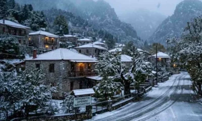 Χιονισμένο ορεινό χωριό με πέτρινα σπίτια και δρόμο καλυμμένο με χιόνι υπό χιονόπτωση.
