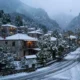 Χιονισμένο ορεινό χωριό με πέτρινα σπίτια και δρόμο καλυμμένο με χιόνι υπό χιονόπτωση.