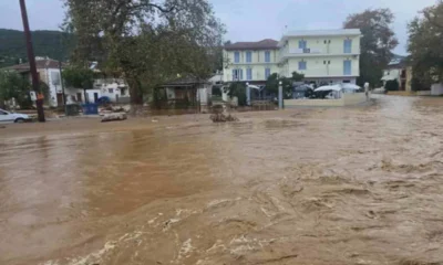 Ορμητικά λασπώδη νερά έχουν καλύψει δρόμο μπροστά από κτίρια και δέντρα.