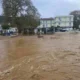 Ορμητικά λασπώδη νερά έχουν καλύψει δρόμο μπροστά από κτίρια και δέντρα.