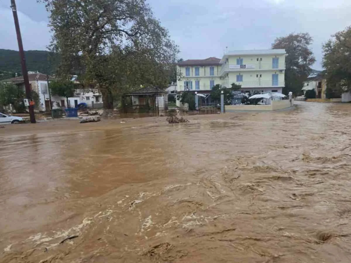 Ορμητικά λασπώδη νερά έχουν καλύψει δρόμο μπροστά από κτίρια και δέντρα.